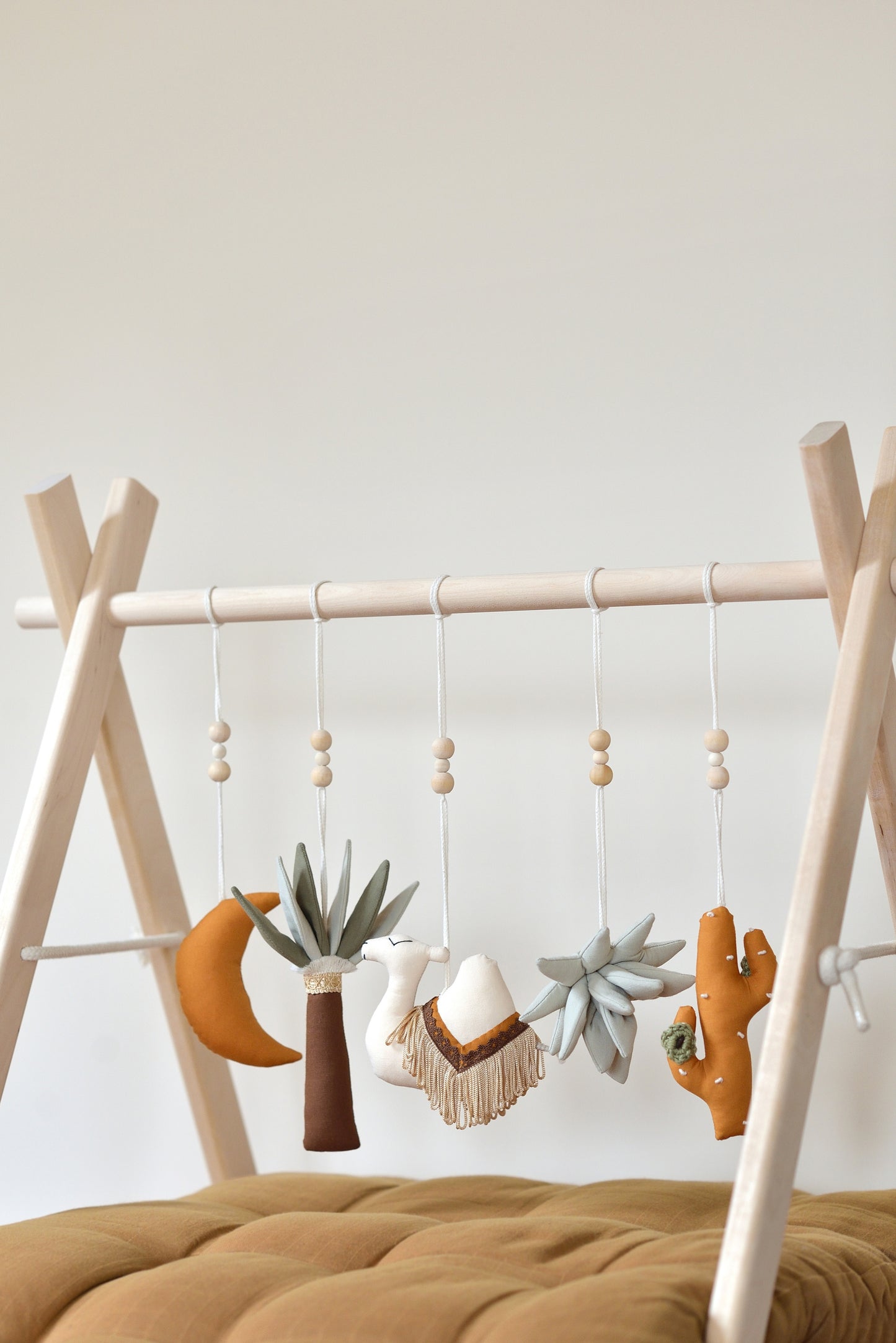 Desert themed play gym toys hanging on a wodden play gym stand: moon, palm, succulent, camel and cacti in khaki, sage, brown and ochre colours. Brown playmat under.