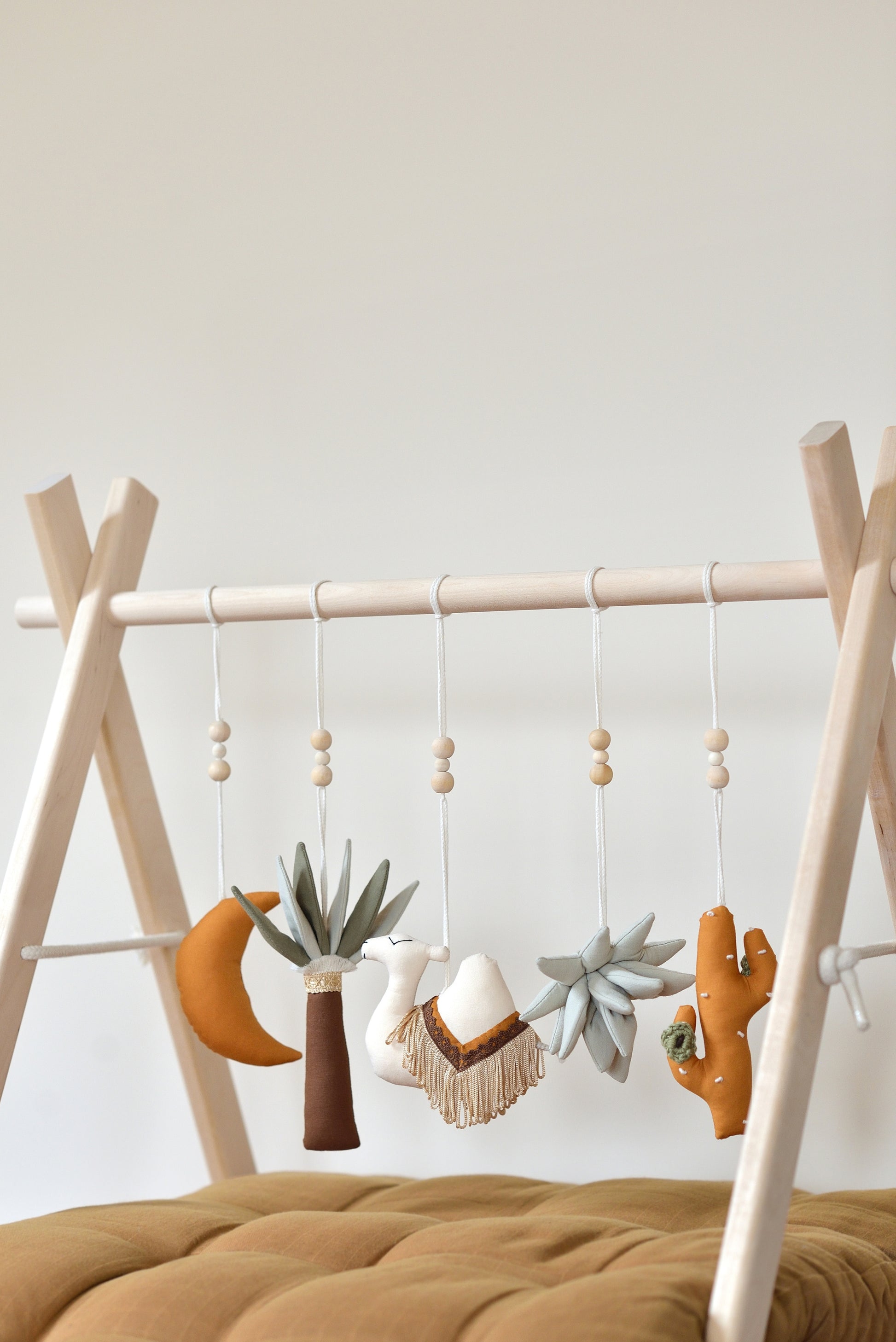 Desert themed play gym toys hanging on a wodden play gym stand: moon, palm, succulent, camel and cacti in khaki, sage, brown and ochre colours. Brown playmat under.