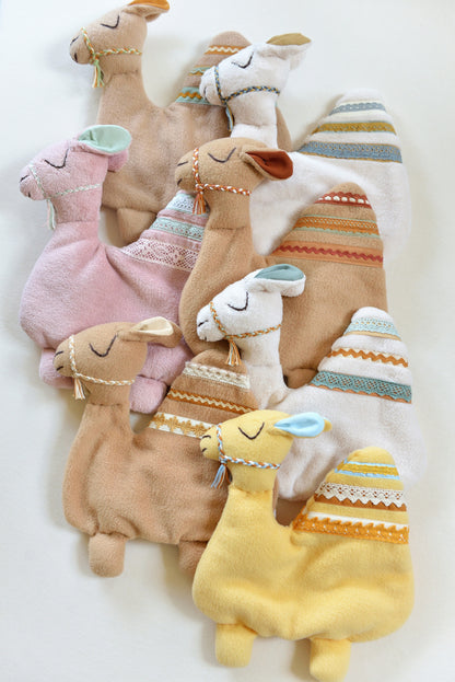 Selection of handmade camel shaped baby comforters with lace details on an ivory background in different colours: ivory, tan, blush pink and yellow.