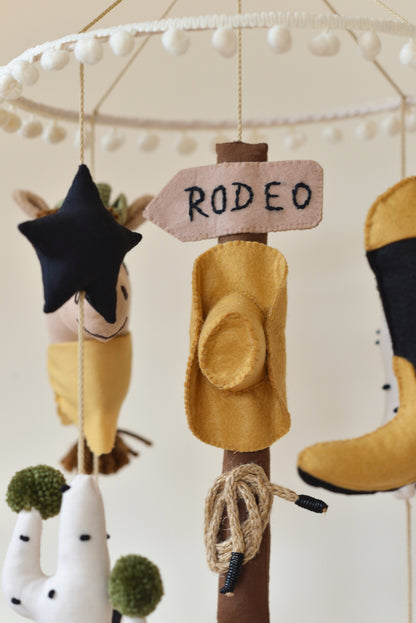Handmade Wild West rodeo nursery mobile with cowboy boots, hats, horses, cacti, stars, and rodeo signpost in boho desert style.