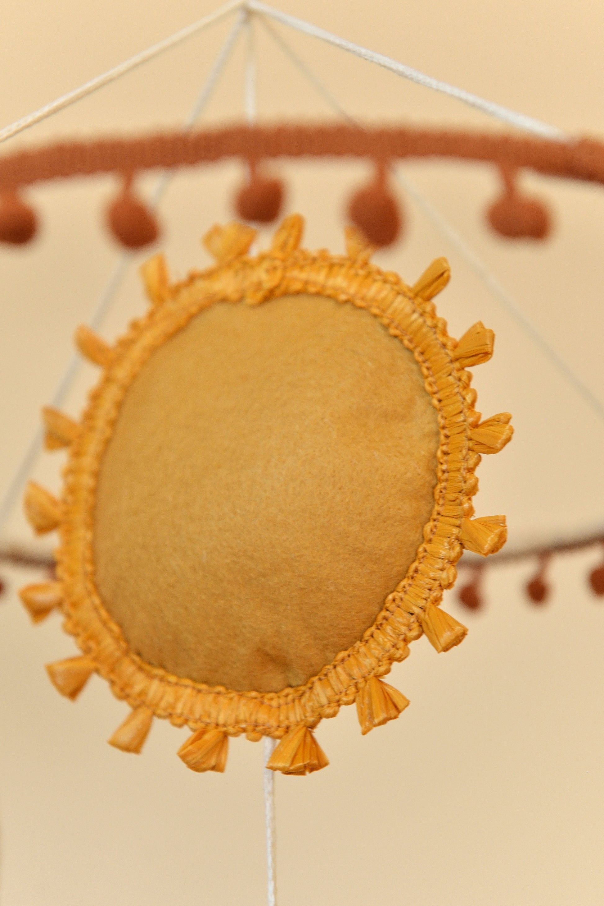 Close up of sun figure on a boho nursery mobile with pom pom trim hanging on the hoop blurred in the foreground.