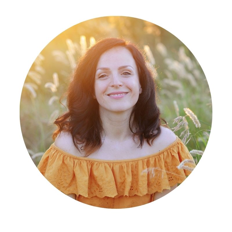 A circular portrait of a smiling woman with dark brown hair wearing an off-the-shoulder marigold blouse, set against a blurred field of wild flowers with warm sunlight.