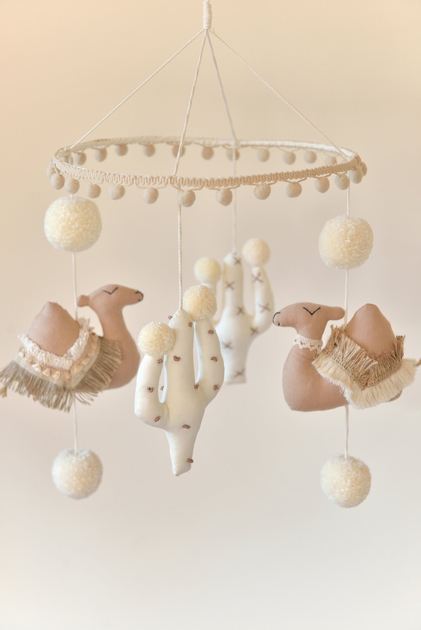 A soft, neutral-colored desert themed boho baby mobile features tan camels, offwhite cacti and midi sized handmade ivory pom poms, hanging from a hoop adorned with light tan pom pom trim, creating a whimsical, calming ambiance.