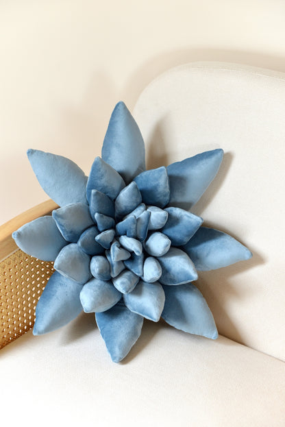 Large succulent shaped velvet decor pillow in sky blue colour placed on an ivory rattan armchair.