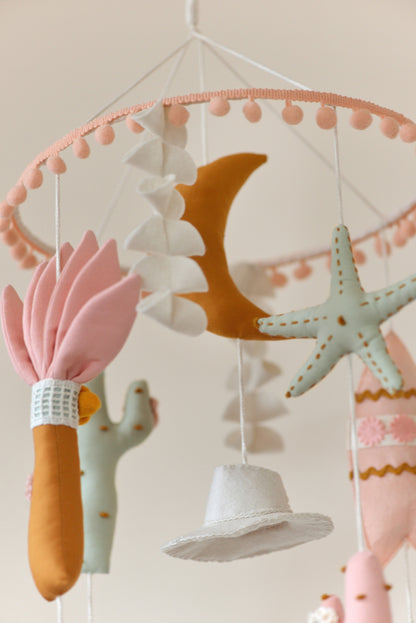 Baby nursery mobile with colorful hanging figures against a light background: moon, palm, starfish, cowboy hat, surf board.