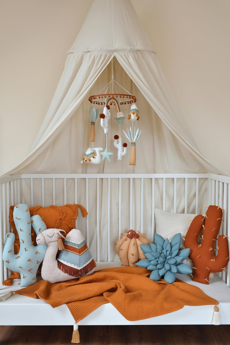 Nursery scene with Morocco Magic Nursery Mobile above a lowered crib. The crib has a cozy ivory canopy hung above and boho cactus pillows  are placed nicely in the crib with a rust blanket under.