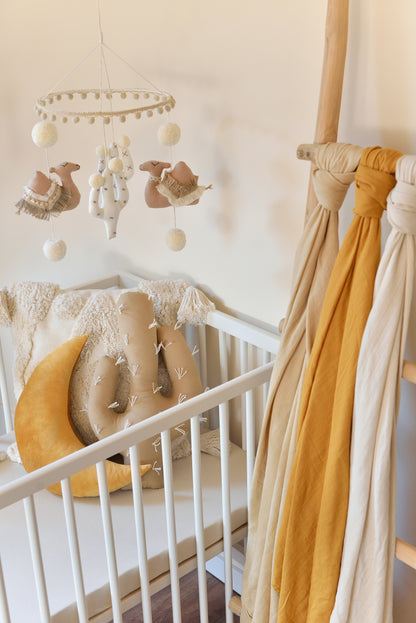 A soft, neutral-colored desert themed boho baby mobile features tan camels, offwhite cacti and midi sized handmade ivory pom poms, hanging from a hoop adorned with light tan pom pom trim. It hangs above a white crib with an ivory tufted pillow and a gold moon and cactus pillow combo in the background. A wooden ladder displaying ivory, tan, gold mustard swaddles is placed in the foreground next to the crib.