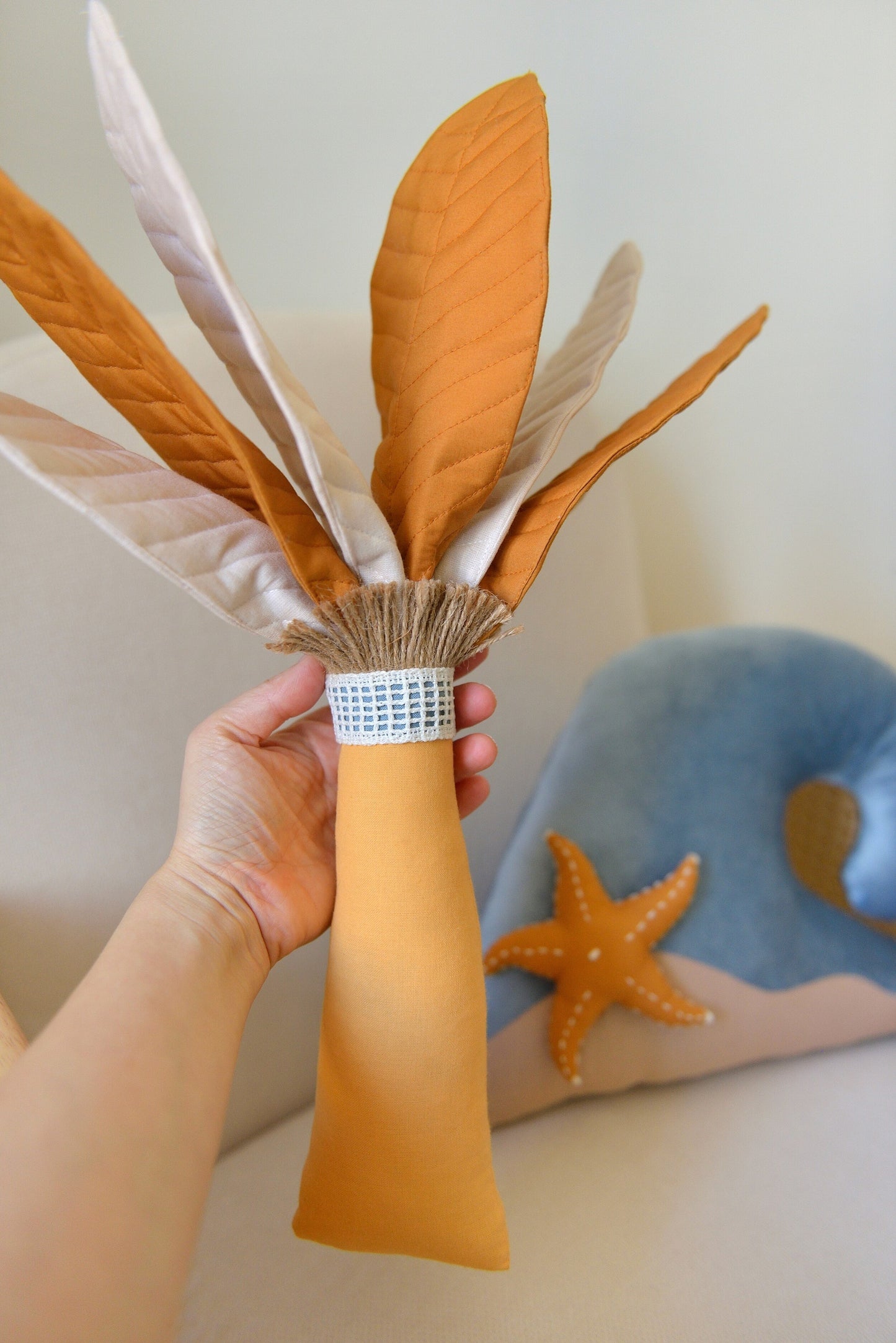 Hand holding Ochre Palm Tree Decor Pillow handmade by Boho Baby Heaven. Featuring mixed ivory and ochre palm fronds and amber trunk. Wave Decor Pillow on a rattan armchair blurred in the background.
