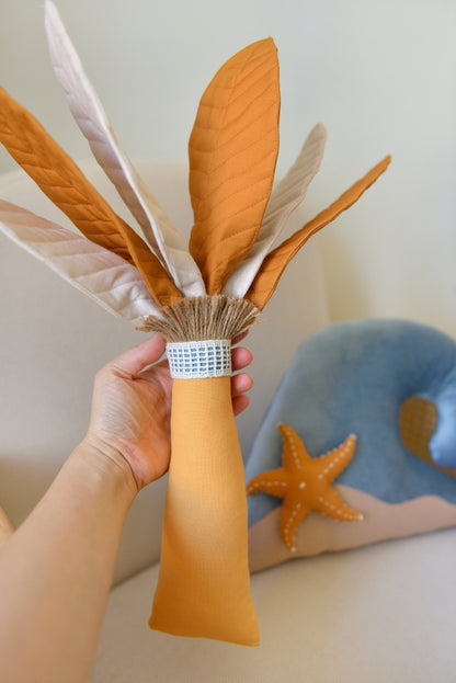 Hand holding Ochre Palm Tree Decor Pillow handmade by Boho Baby Heaven. Featuring mixed ivory and ochre palm fronds and amber trunk. Wave Decor Pillow on a rattan armchair blurred in the background.