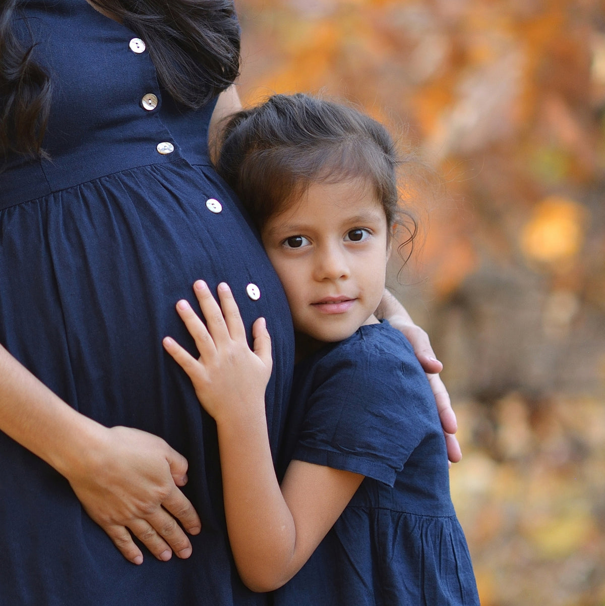 A pregnant woman in a navy maternity gown and is embraced by a little girl standing.