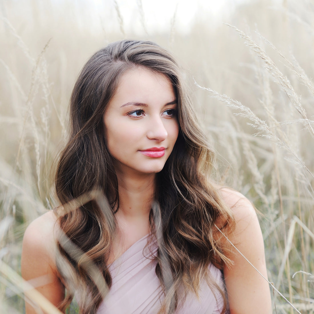 A young woman with long wavy brown hair wearing a light pink one-shoulder top, posing outdoors in a field of dry beige grass.