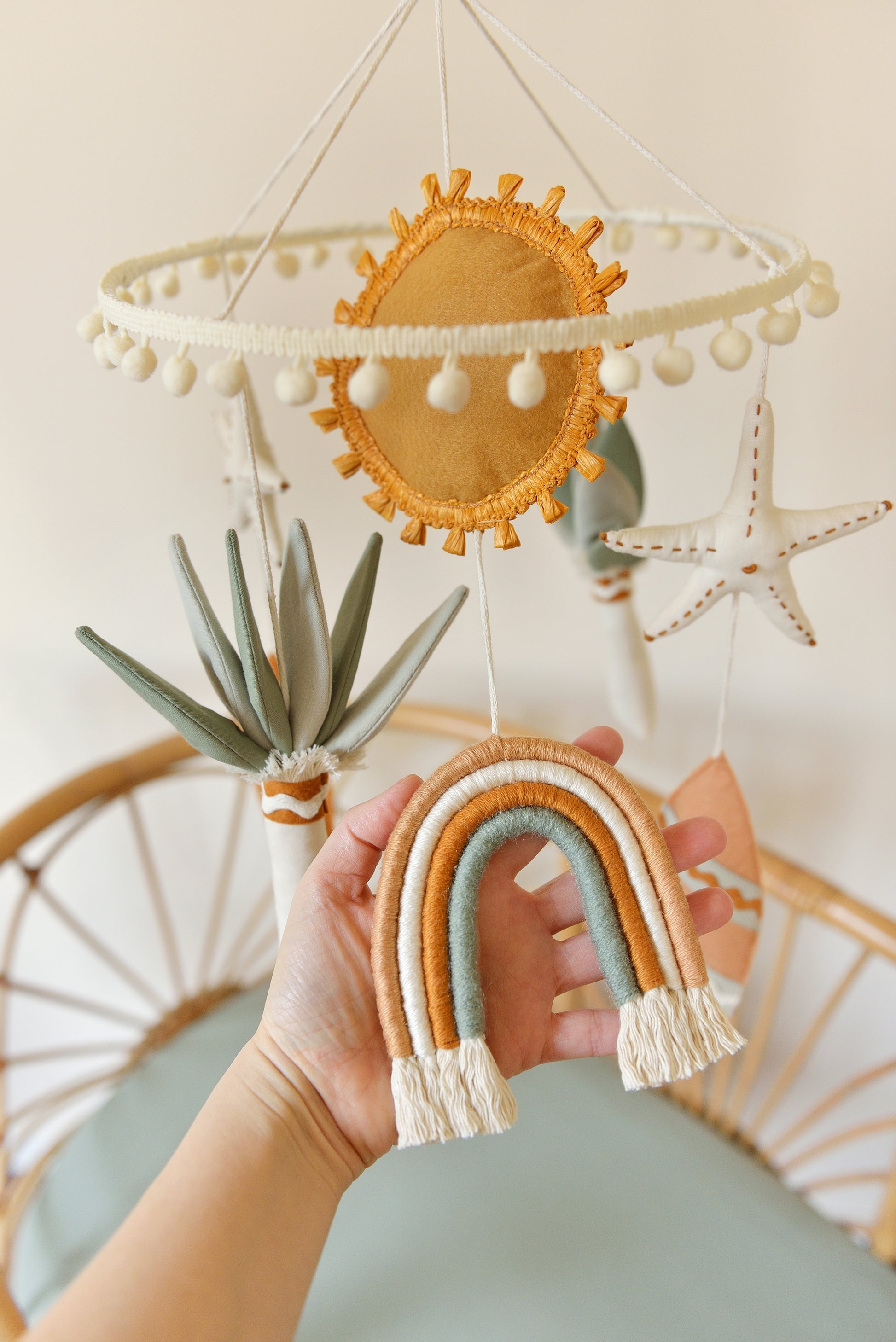 Hand holding a rainbow figure hanging off a nursery mobile hoop. Other figures include sun, starfish, surf board, palm. Mobile is hung over a rattan bassinet.