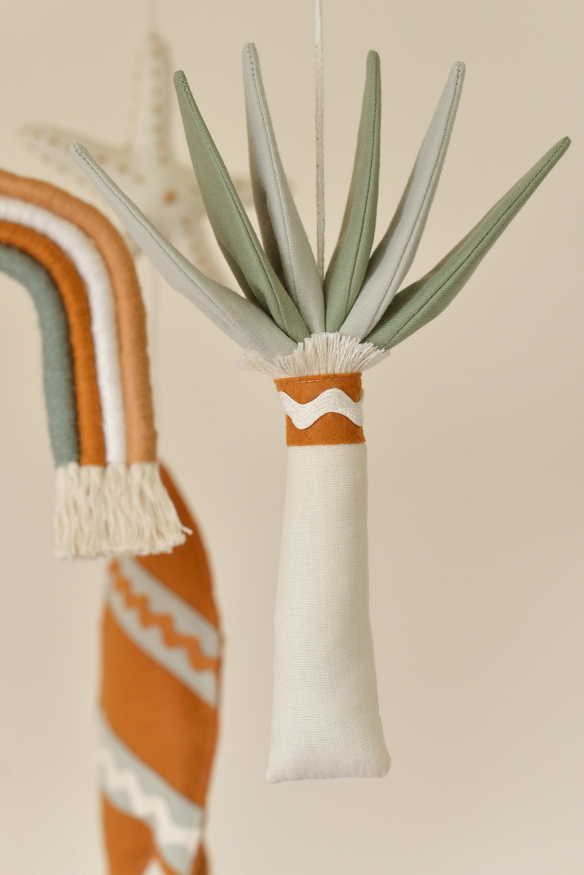 Close up of palm tree figure with ivory trunk and mixed sage leaves on a boho nursery mobile. Rainbow and surf board figures blurred in the background.