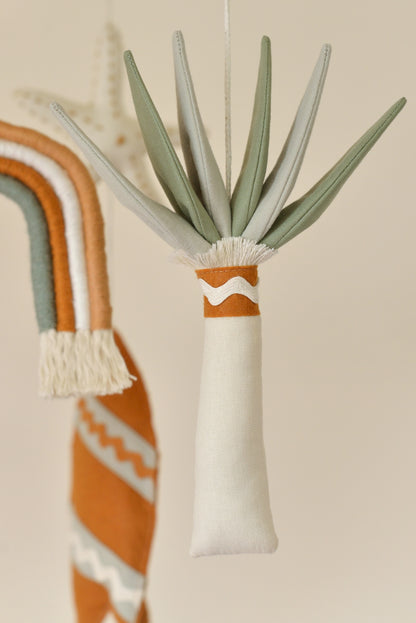 Close up of palm tree figure with ivory trunk and mixed sage leaves on a boho nursery mobile. Rainbow and surf board figures blurred in the background.