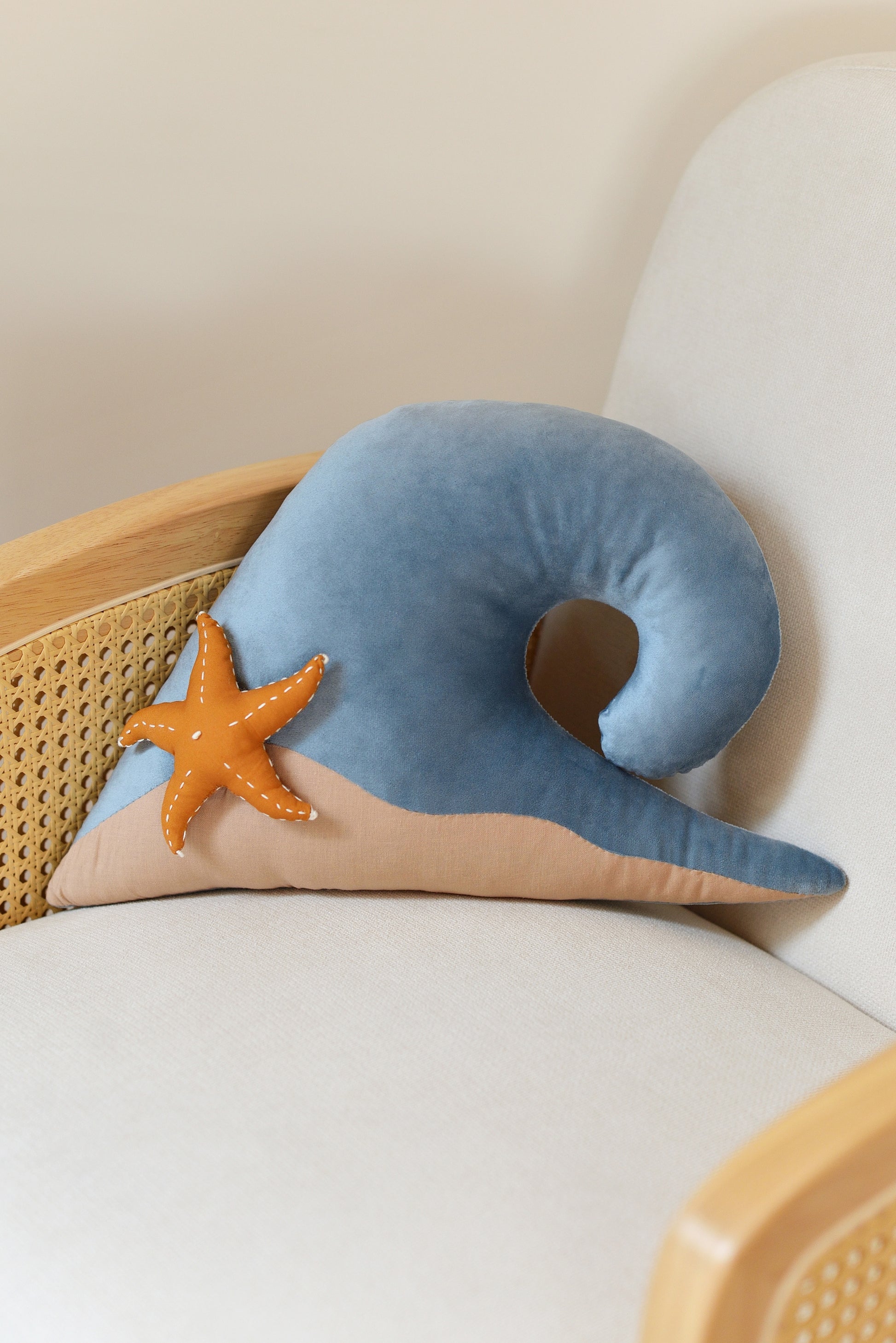 Sky Blue wave shaped decor pillow with tan sandy base and ochre starfish on an ivory rattan armchair.