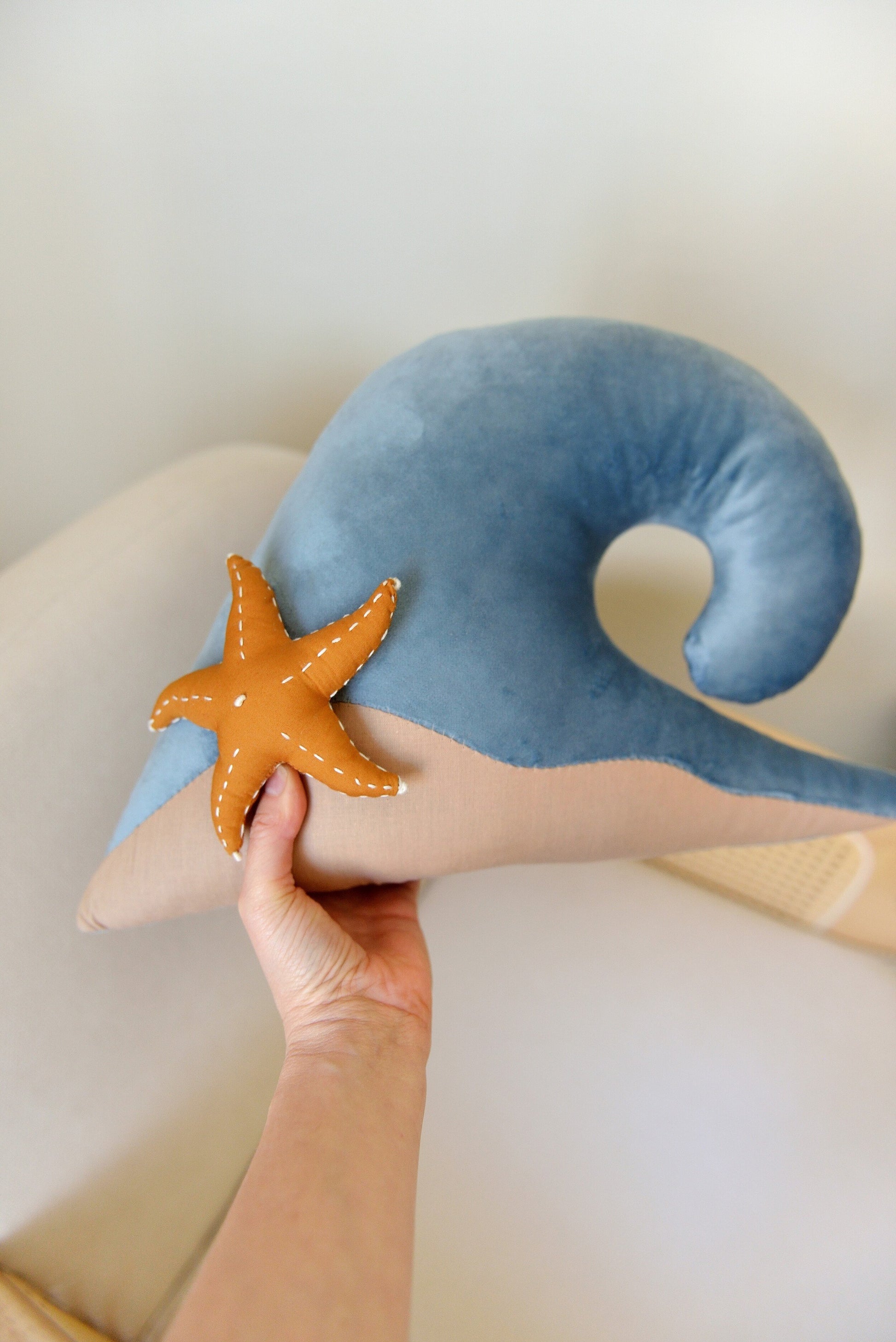 Hand holding an ocean wave shaped decor pillow with a starfish aobve a rattan armchair in front of a white wall.