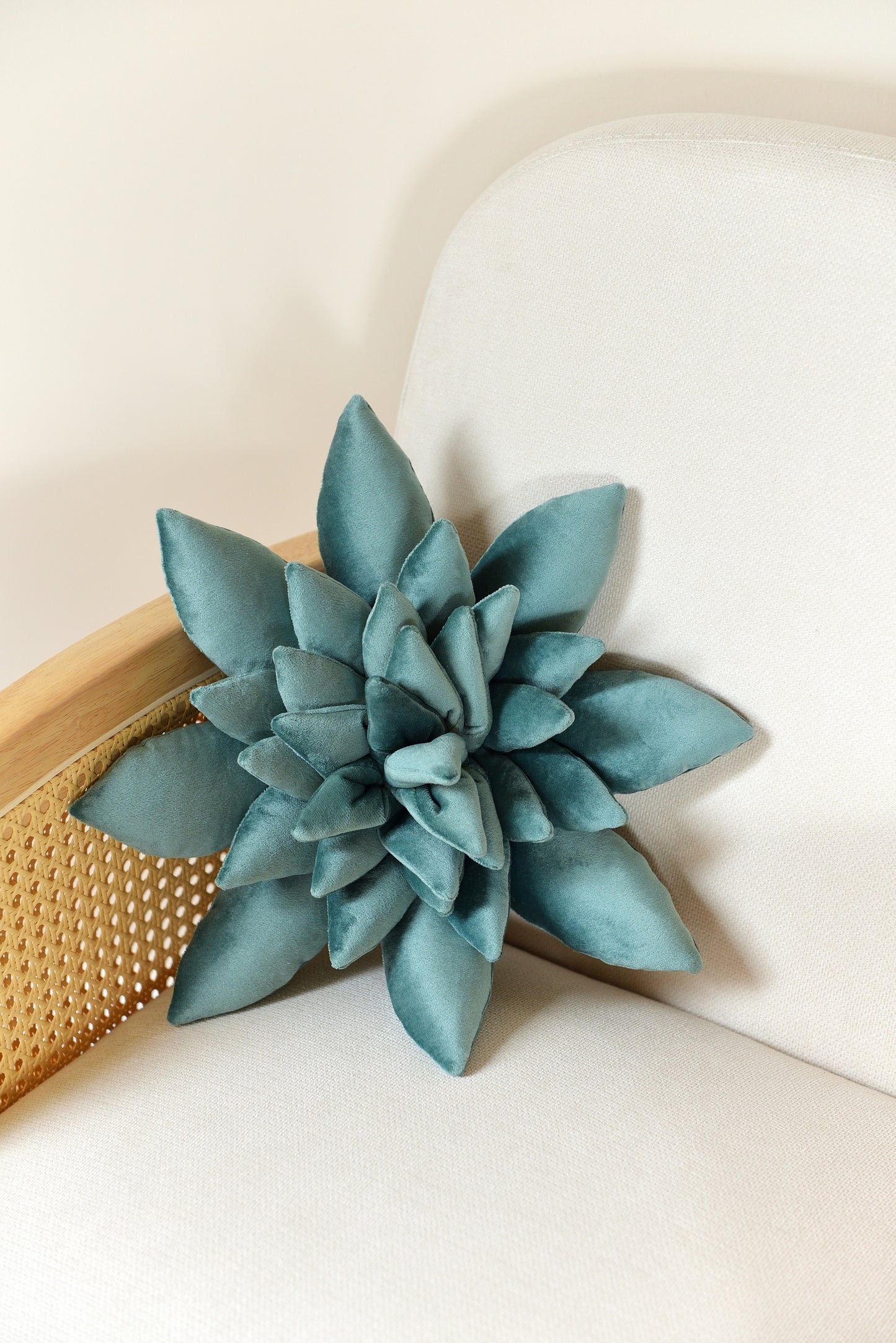 Standard size succulent shaped velvet decor pillow in teal colour placed on an ivory rattan armchair.