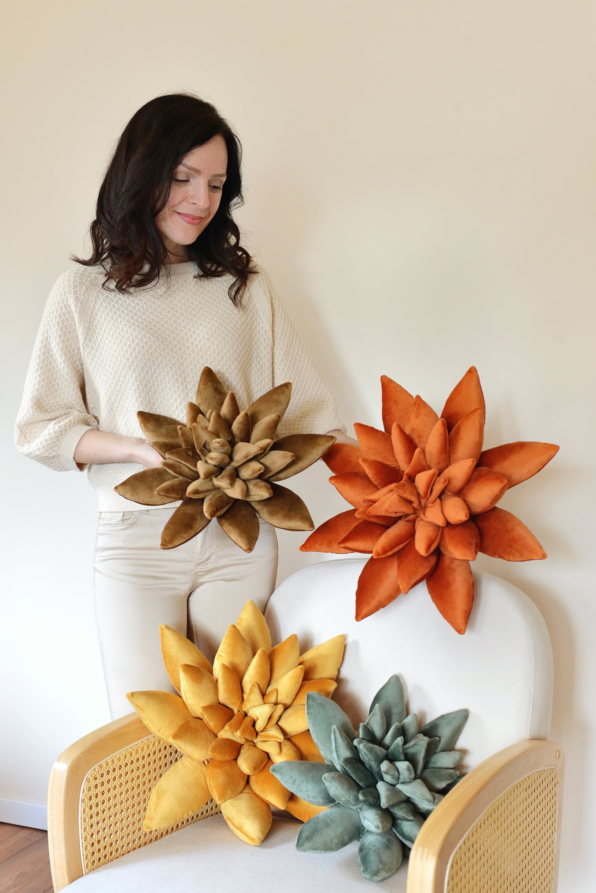 A woman poses with a collection of standard and large size, plush succulent-shaped decor pillows. The pillows are in shades golden yellow, coffee brown, jade green and rust. They are arranged on a white rattan armchair and held by the woman.