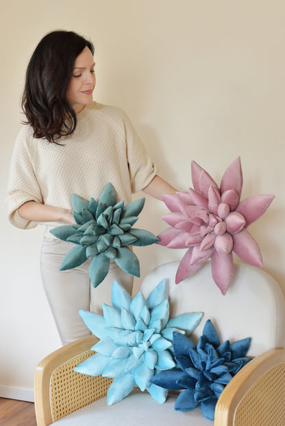 A woman poses with a collection of standard and large size, plush succulent-shaped decor pillows. The pillows are in shades of teal, lilac, aqua, and dark blue. They are arranged on a white rattan armchair and held by the woman.