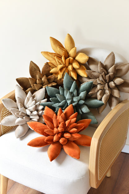 A selection of velvet succulent shaped decor pillows in shades of golden yellow, pearl, sand, coffee brown, jade green and rust. The cushions are arranged on an ivory rattan armchair against a light background.