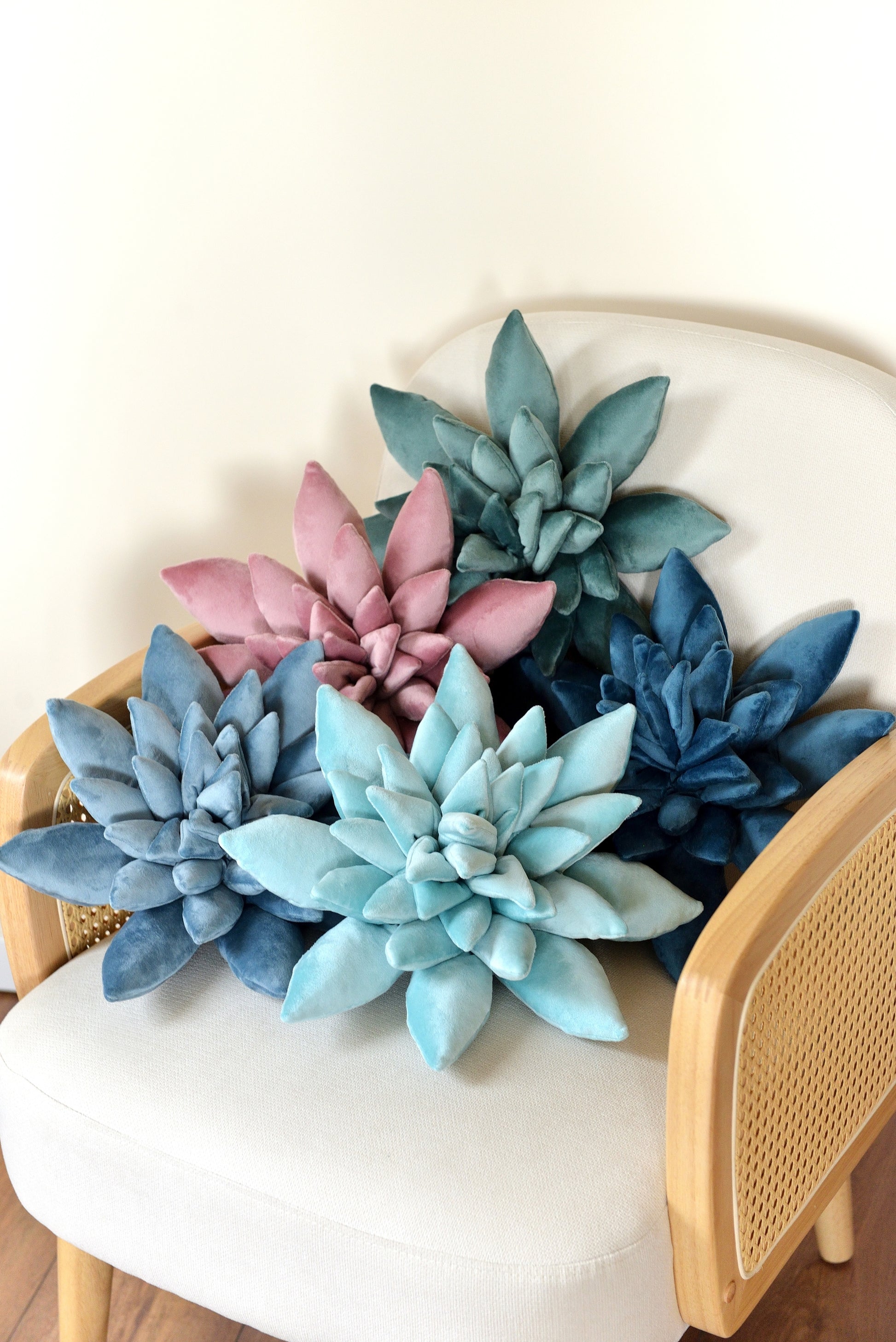 A selection of velvet succulent shaped decor pillows in shades of teal, lilac, aqua, sky and ocean blue. The cushions are arranged on an ivory rattan armchair against a light background.