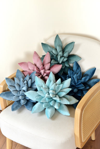 A selection of velvet succulent shaped decor pillows in shades of teal, lilac, aqua, sky and ocean blue. The cushions are arranged on an ivory rattan armchair against a light background.