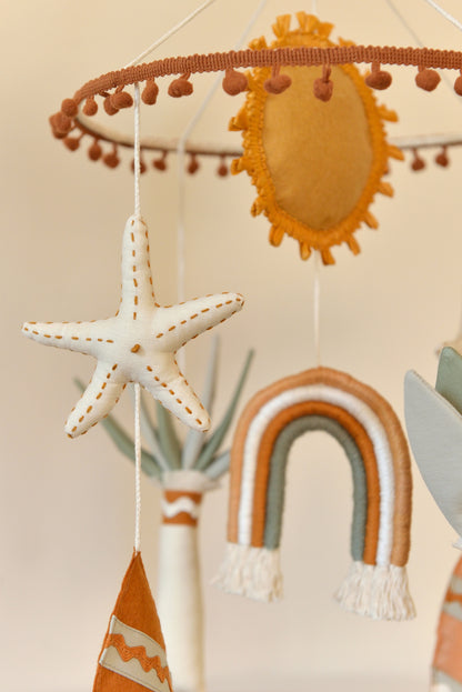 Close up of an ivory starfish figure hanging on a boho nursery mobile. Rainbow, sun, palm tree and surf board figures blurred in the background.