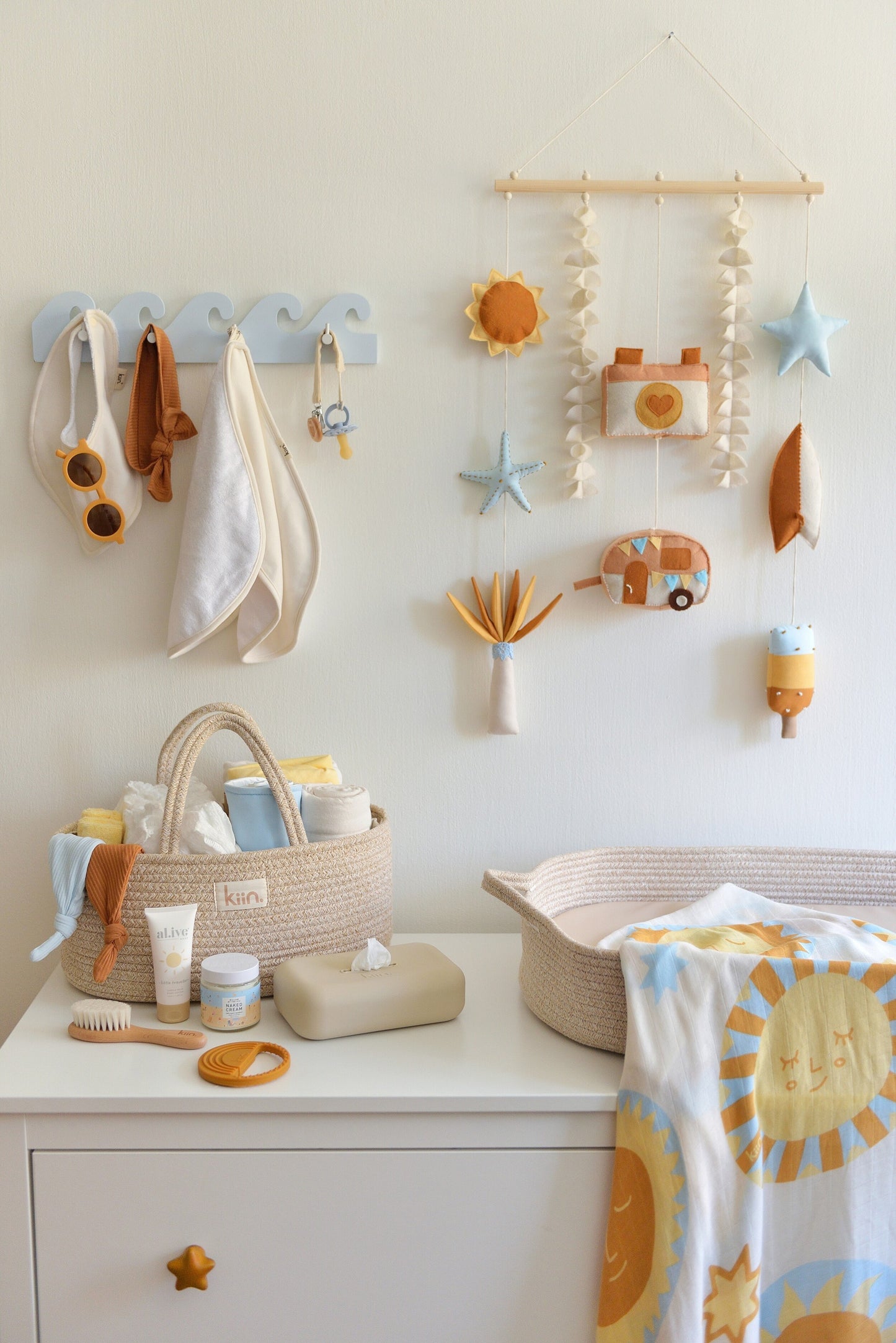 Nappy changing station with baby items  on a white dresser. Surf-themed wall hanging and wave-shaped wall hanger above the dresser. Nappy caddy and nappy changing basket with sun themed swaddle on top of the dresser.