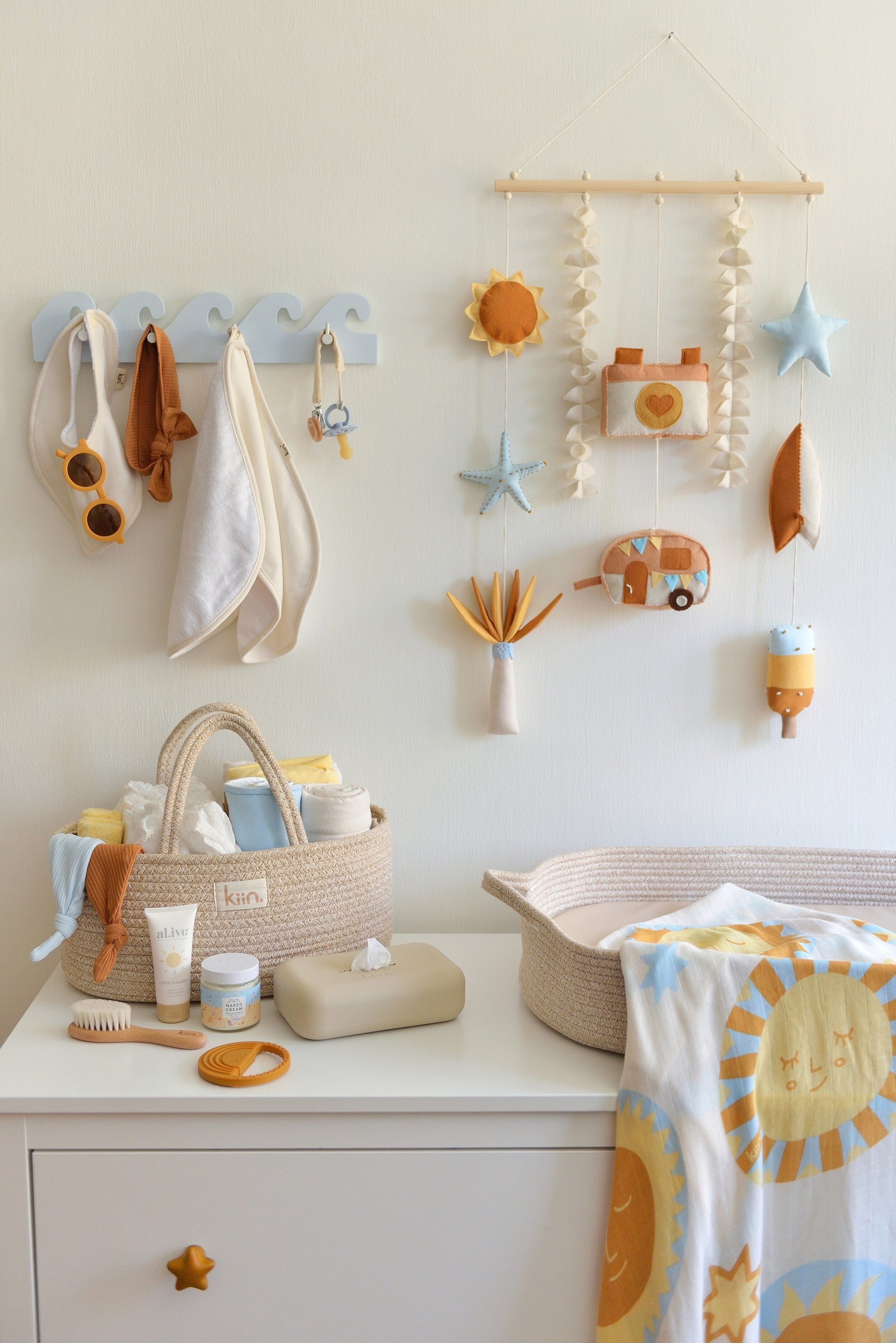 Nappy changing station with baby items  on a white dresser. Surf-themed wall hanging and wave-shaped wall hanger above the dresser. Nappy caddy and nappy changing basket with sun themed swaddle on top of the dresser.