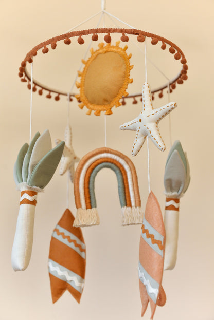 Handmade boho nursery mobile with rusty surf boards, ochre sun, rainbow and sage palm tree figures hanging off a hoop adorned with rust coloured mini pom pom trim.