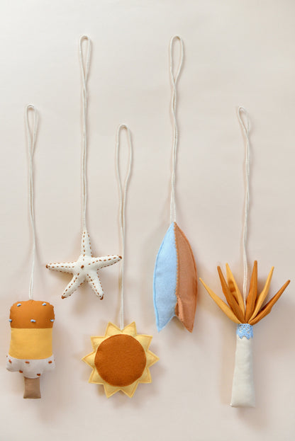 Flatlay of handmade hanging baby play gym toys against a light background: popsickle, surf board, sun, starfish, palm tree.