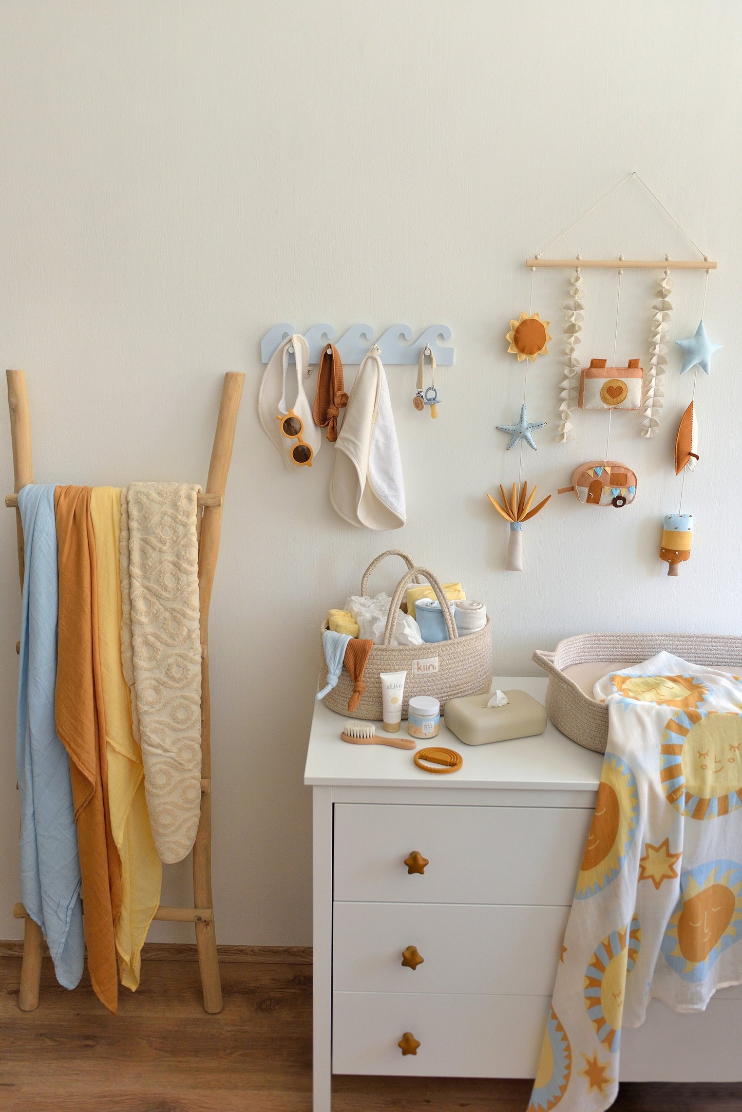 Nappy changing station with baby items  on a white dresser. Surf-themed wall hanging and wave-shaped wall hanger above the dresser, wooden ladder with swaddles displayed on the left.