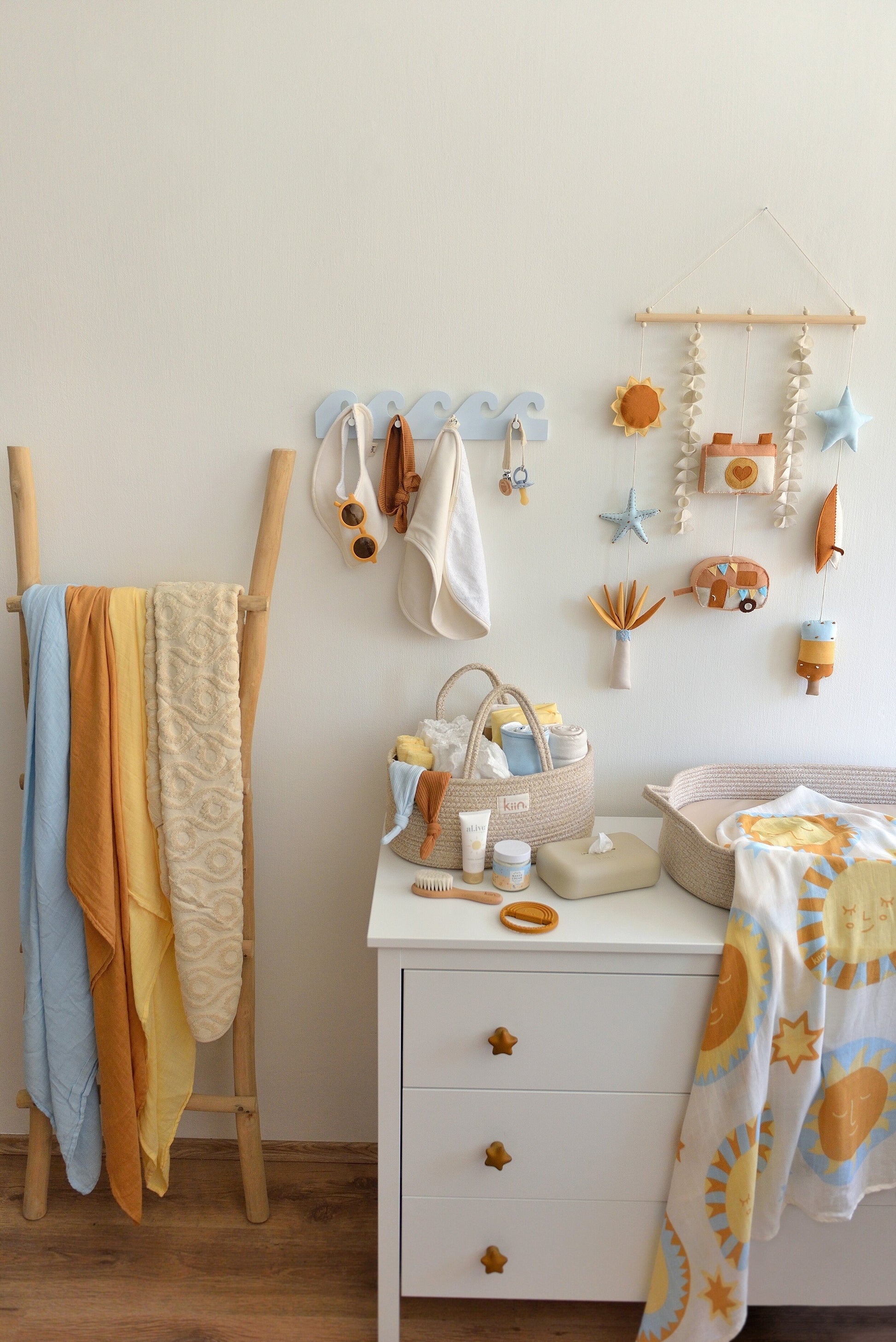 Nappy changing station with baby items  on a white dresser. Surf-themed wall hanging and wave-shaped wall hanger above the dresser, wooden ladder with swaddles displayed on the left.