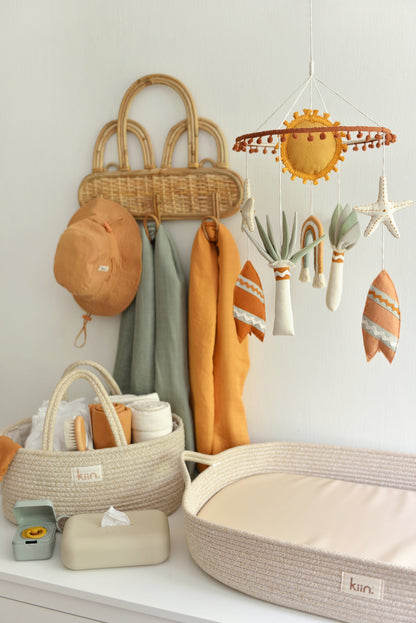 Sun, surf and rainbow themed nappy chaging area for baby in gender neutral style. Features coton rope changing basket, nappy caddy on a white dresser with baby essentials in ivory, rust and sage shades. Rattan wall hanger holds swaddles and baby hat. Nursery mobile featuring sun, rusty surf boards, ivory starfish and sage palms hangs above the changing basket.  