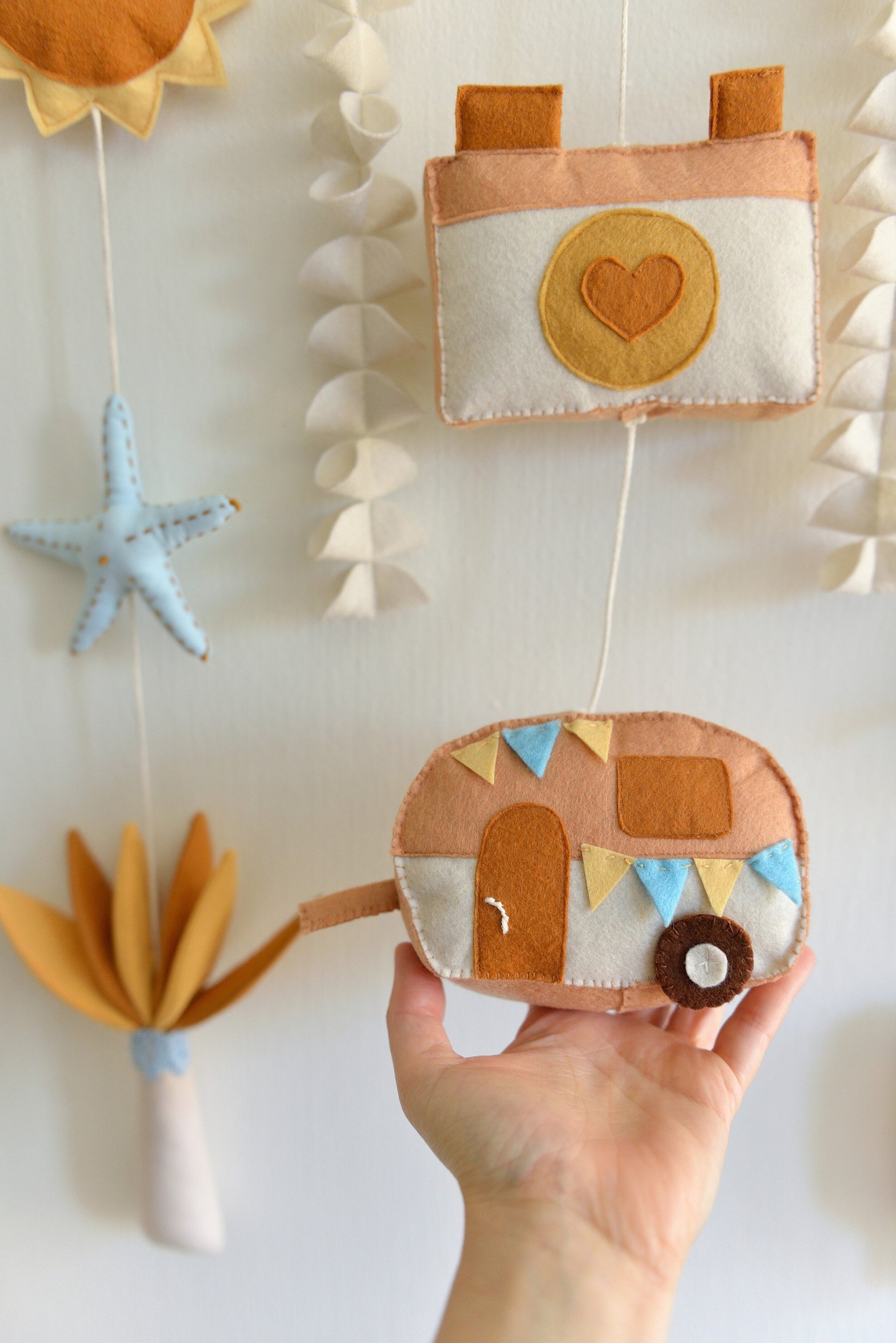 Hand holding a felt camper figure  against a white wall. Other felt figures of the rod hanging bunting are blurred in the background: camera, palm tree, starfish, sun, seaweed.