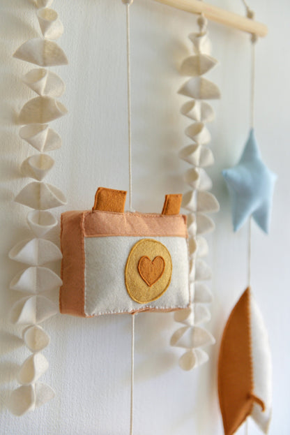 Camera figure up close against a white wall. Other felt figures of the rod hanging bunting are blurred in the background: star, surf board, seaweed.