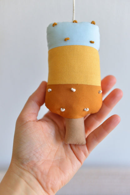 Hand holding a small popsicle figure with a light blue top, amber middle and ochre bottom.