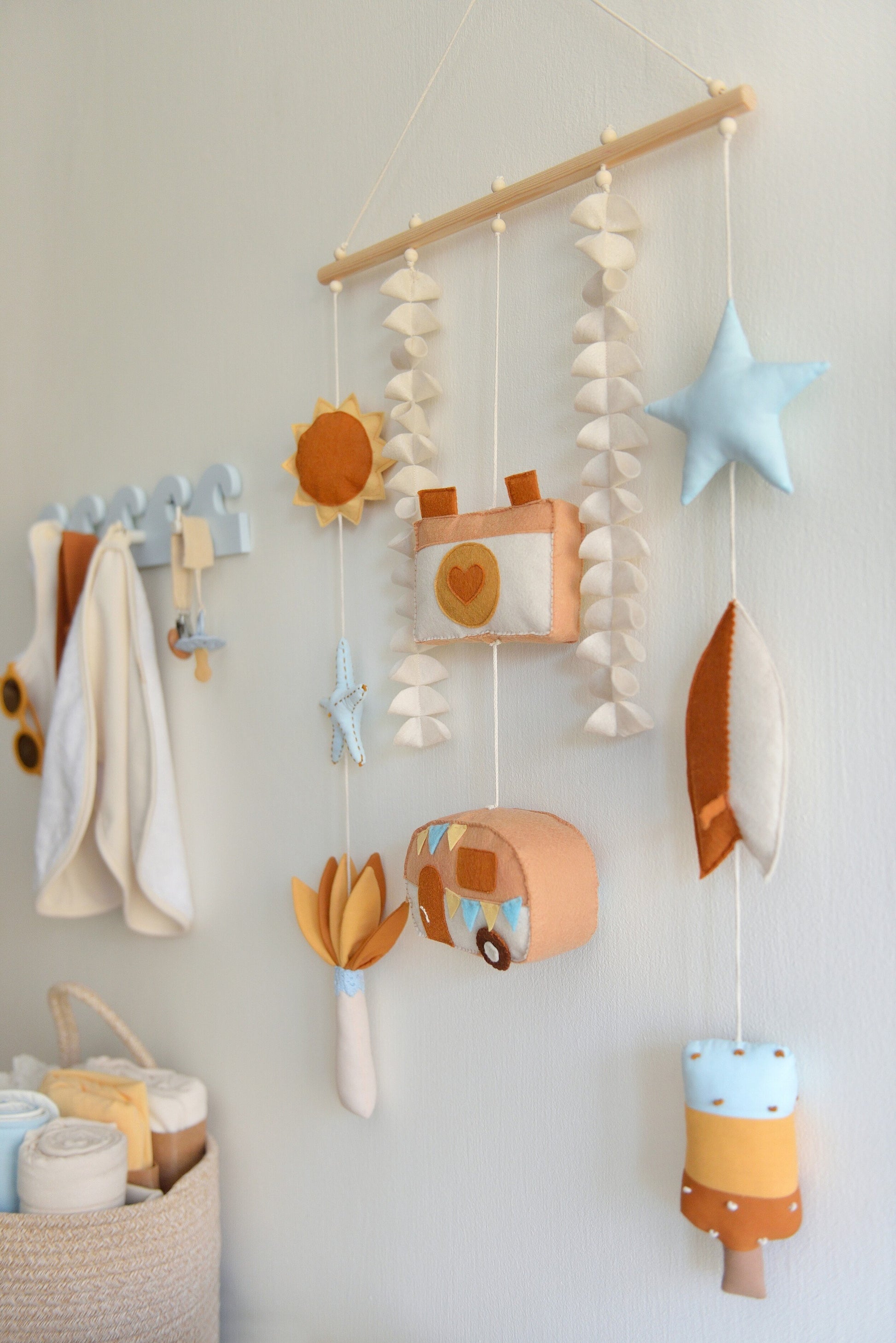 Decorative wall hanging (rod-Hanging Bunting) with colorful felt shapes against a white background. Figures hang off a wooden rod: sun, starfish, palm tree, camera, camper van, star, surf board, popsicle. Other baby essentials blurred in the background.