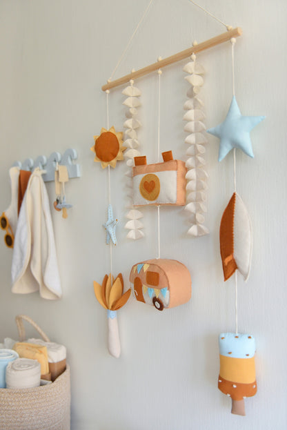Decorative wall hanging (rod-Hanging Bunting) with colorful felt shapes against a white background. Figures hang off a wooden rod: sun, starfish, palm tree, camera, camper van, star, surf board, popsicle. Other baby essentials blurred in the background.