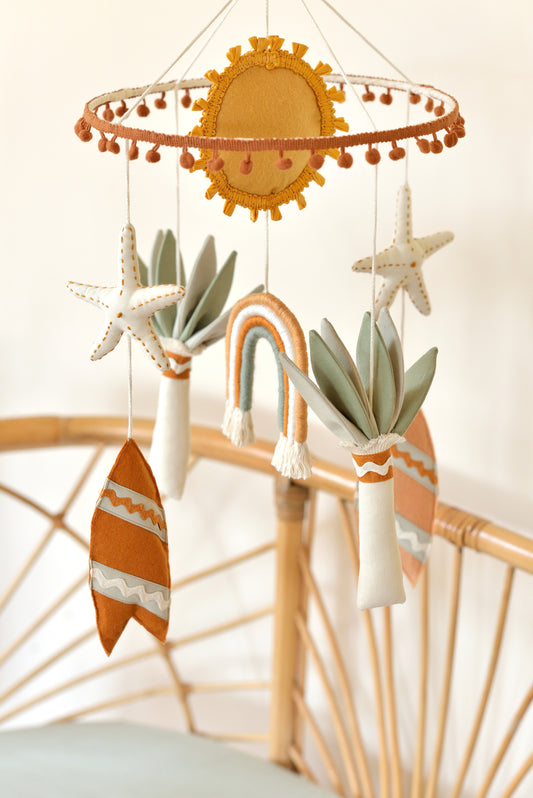 Handmade boho nursery mobile with rusty surf boards, ochre sun, rainbow and sage palm tree figures hanging off a hoop adorned with rust coloured mini pom pom trim. Rattan bassinet with sage mattress blurred in the background.