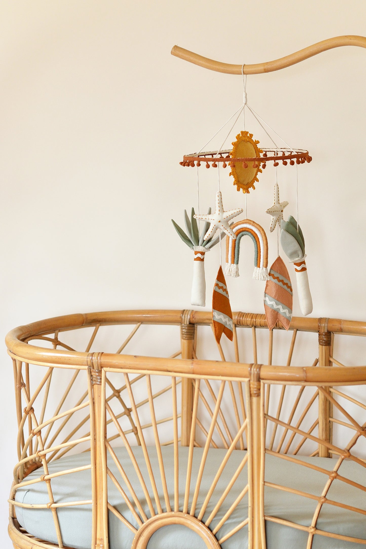 Handmade boho nursery mobile with rusty surf boards, ochre sun, rainbow and sage palm tree figures hanging off a hoop adorned with rust coloured mini pom pom trim. The mobile hangs above a rattan bassinet with sage mattress.