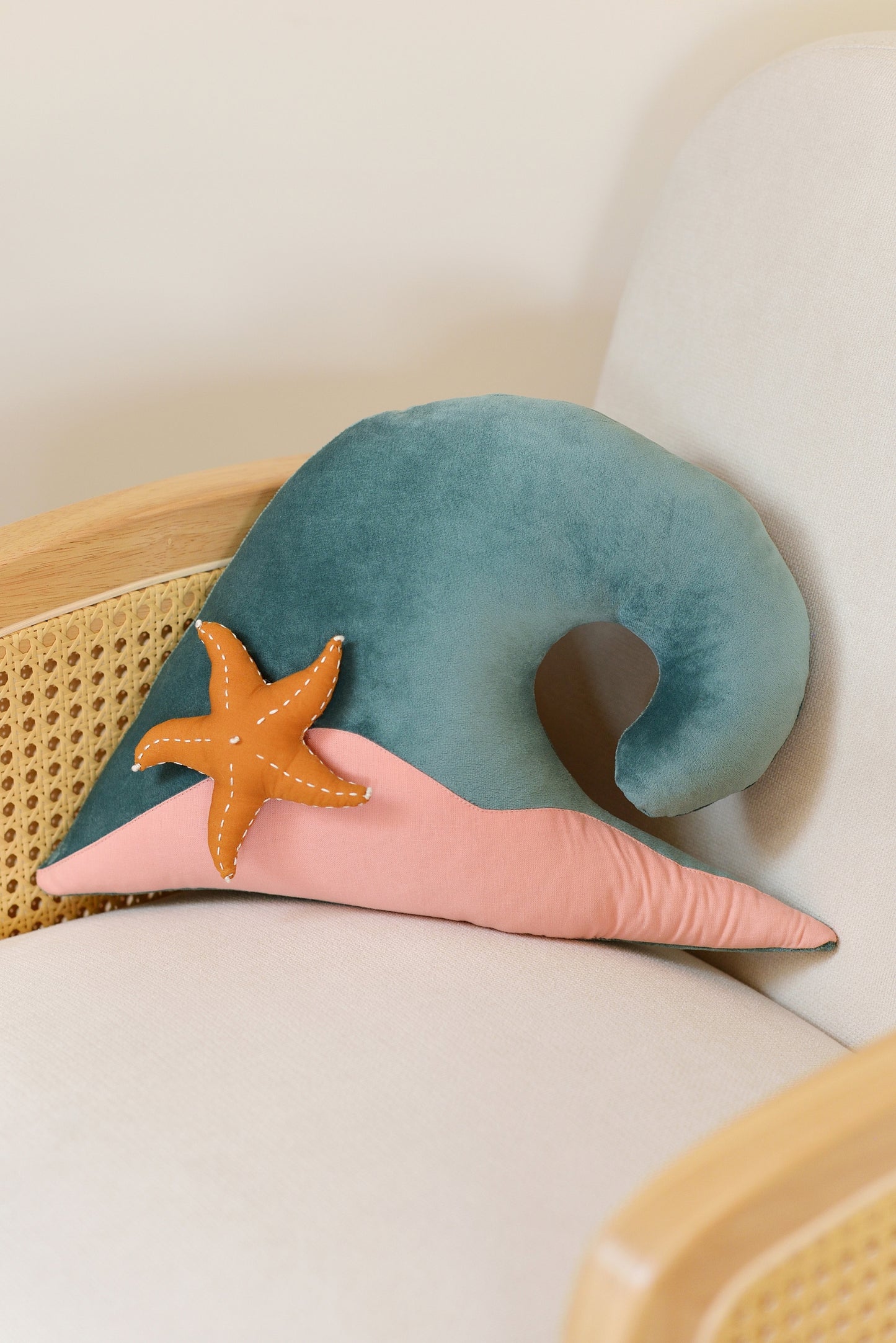 Teal wave shaped decor pillow with pastel pink sandy base and ochre starfish on an ivory rattan armchair.