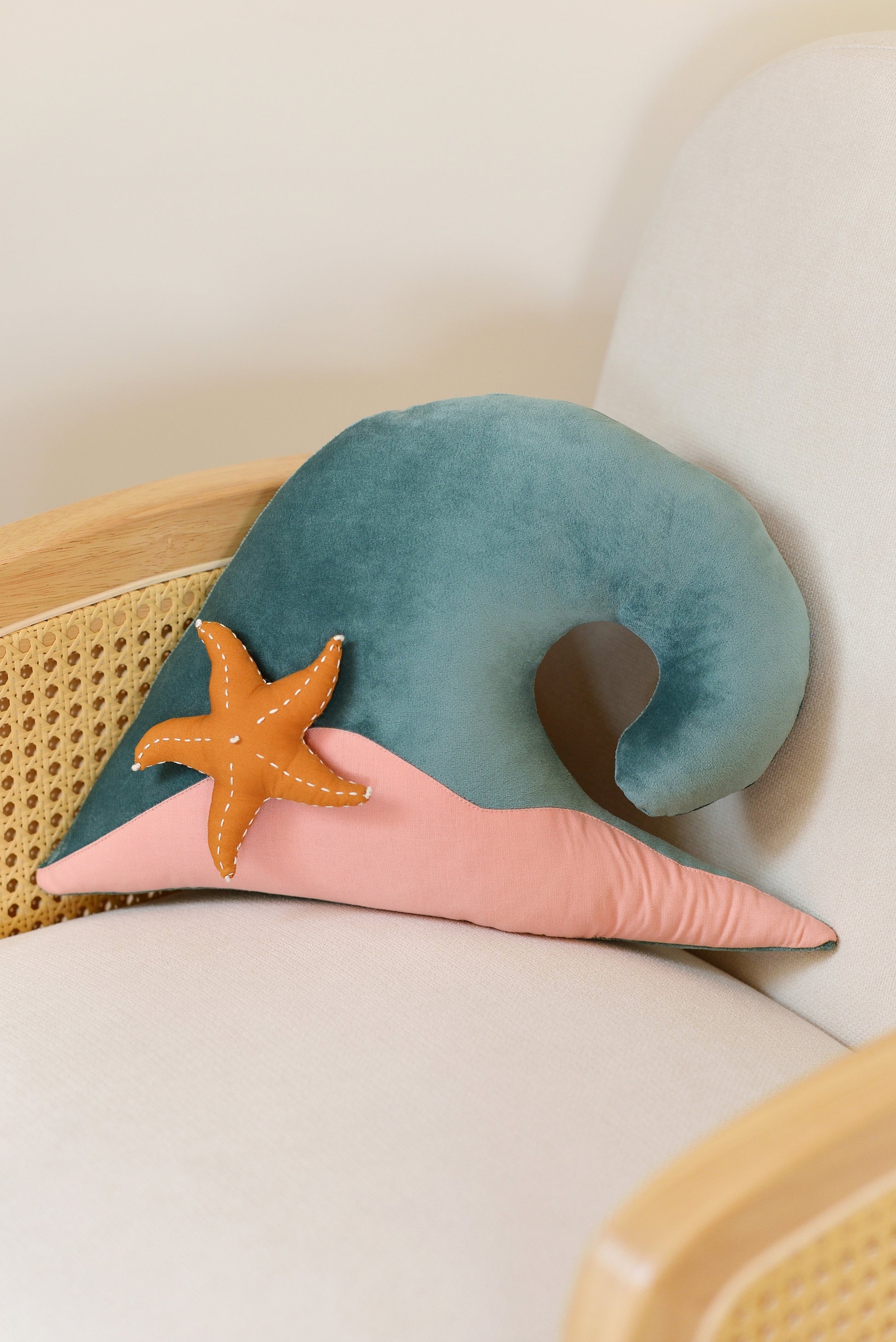 Teal wave shaped decor pillow with pastel pink sandy base and ochre starfish on an ivory rattan armchair.