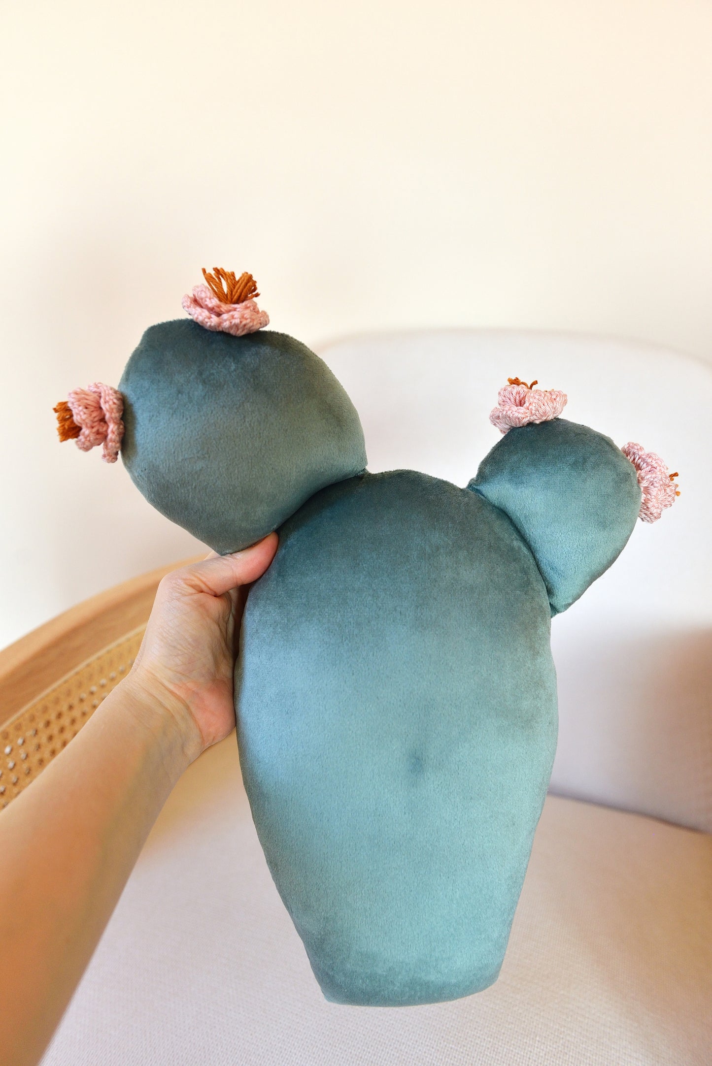 Hand holding prickly pear shaped cactus decor pillow in front of an armchair.