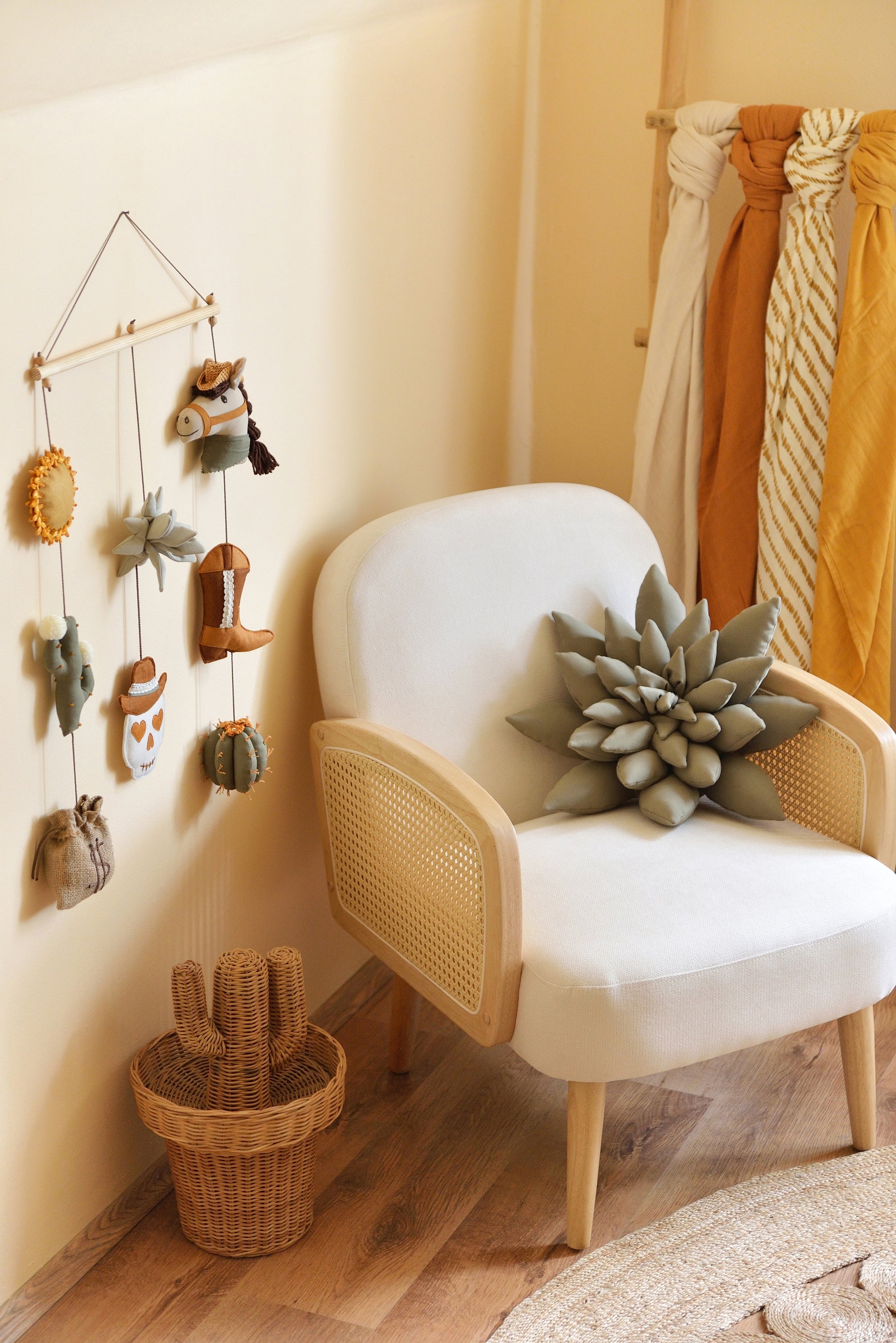 Ivory attan armchair with succulent pillow and a cowboy-themed rod-hanging bunting in a baby room. Woven rattan cactus shaped basket next to it and wooden ladder with colourful swaddles in the background.