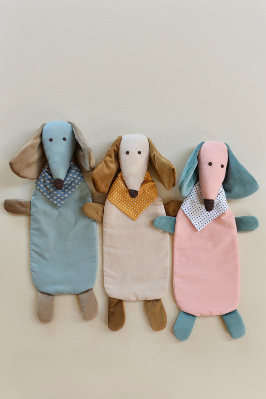 Three Dachshund Comforters with long ears and snouts in blue, cream, and pink. Each wears a patterned bandana, conveying a playful and cozy feel.