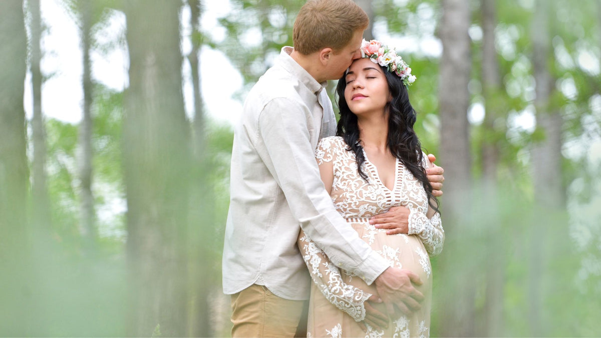 A couple embracing in a forest, with the pregnant woman wearing a cream lace maternity gown and a floral crown, and the man in a light beige shirt and khaki pants.